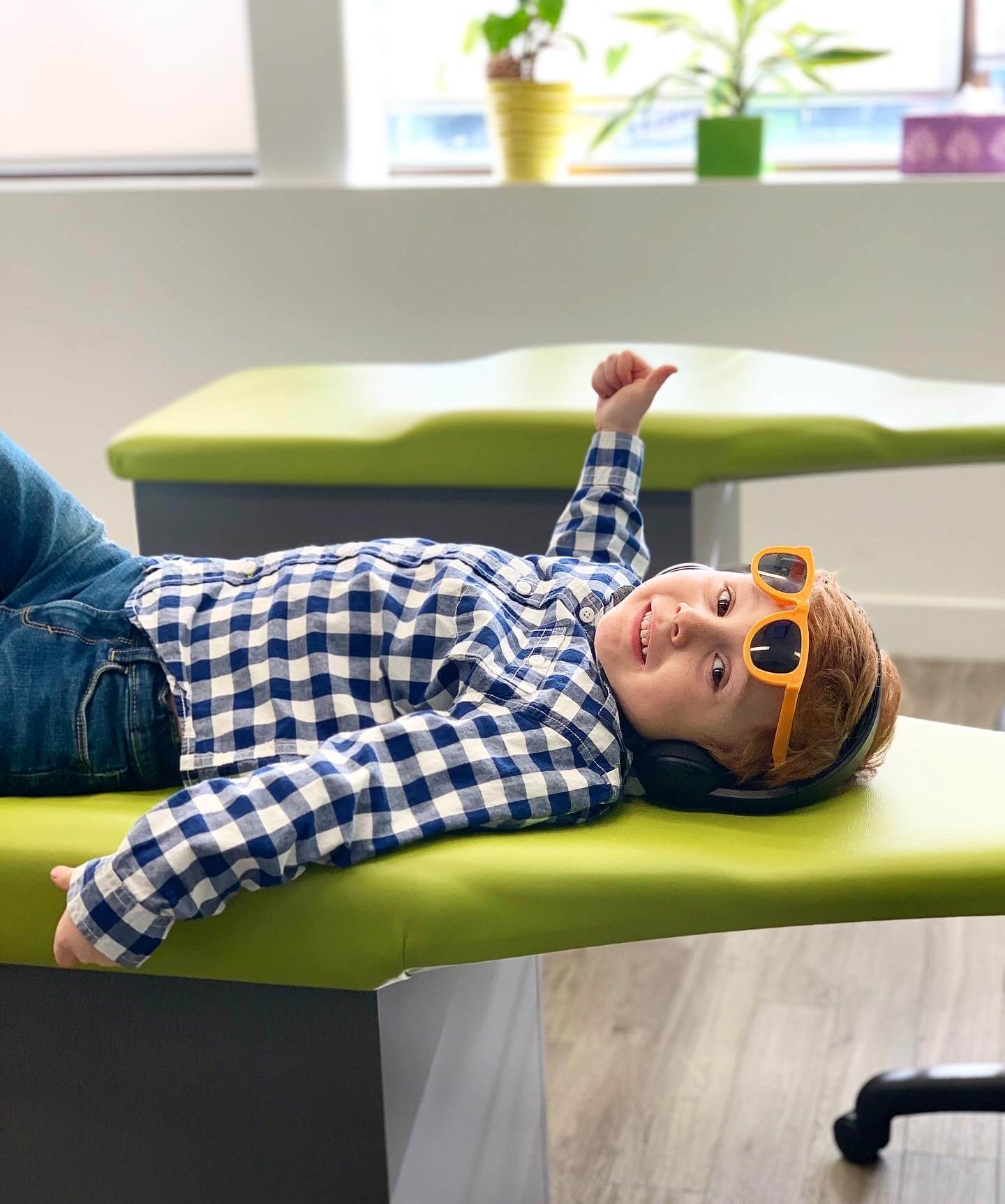 A young boy smiling, giving a thumbs up while lying on a patient's chair.
